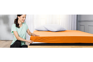 Eine junge Frau in einem grünen T-Shirt bezieht ein grosses Bett mit einem orangen Fixleintuch. Auf dem Bett liegen zwei weisse Kissen. Im Hintergrund scheint Licht durch weisse Vorhänge. 
