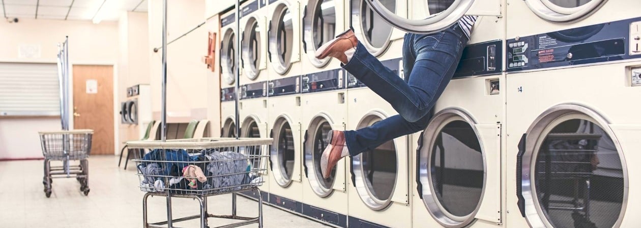 Eine Frau lehnt sich in eine Waschmaschine in einem Waschsalon. Man sieht nur ihre Beine aus der Waschmaschine hängen. In einem Korb hinter ihr ist farbige Wäsche. 